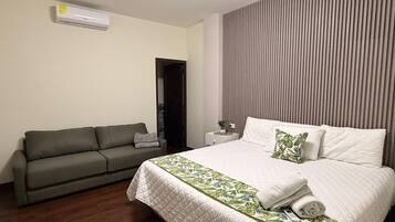A hotel room with a bed, a grey sofa, a small bedside table with a plant, and a wall-mounted air conditioner.