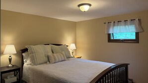 Un dormitorio con una cama, dos mesitas de noche con lámparas, un ventanal con cortinero y una lámpara en el techo.