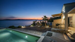 Zona de piscina exterior moderna amb vista al mar al capvespre, un patí amb seients i una zona de menjador amb envans de vidre.