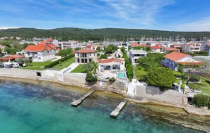 Une zone résidentielle côtière, avec des maisons, une mer d’un bleu limpide et un paysage vallonné en arrière-plan.