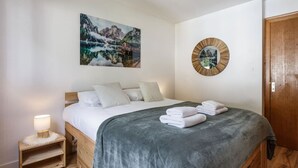 A bedroom with a bed, pillows, a bedside table with a lamp, a wall-mounted mirror, and a painting of a mountain landscape.