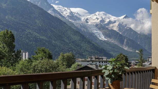A mountain view with snow-capped peaks, a balcony with a potted plant, and buildings in the foreground.