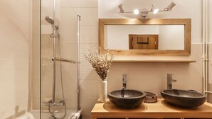 A modern bathroom with a glass-enclosed shower, two vessel sinks, a wooden vanity, a mirror, and a vase with dried flowers.