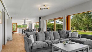 Ein modernes Wohnzimmer mit einer grauen Eckcouch, einem Holztisch und großen Fenstern mit Blick ins Freie.