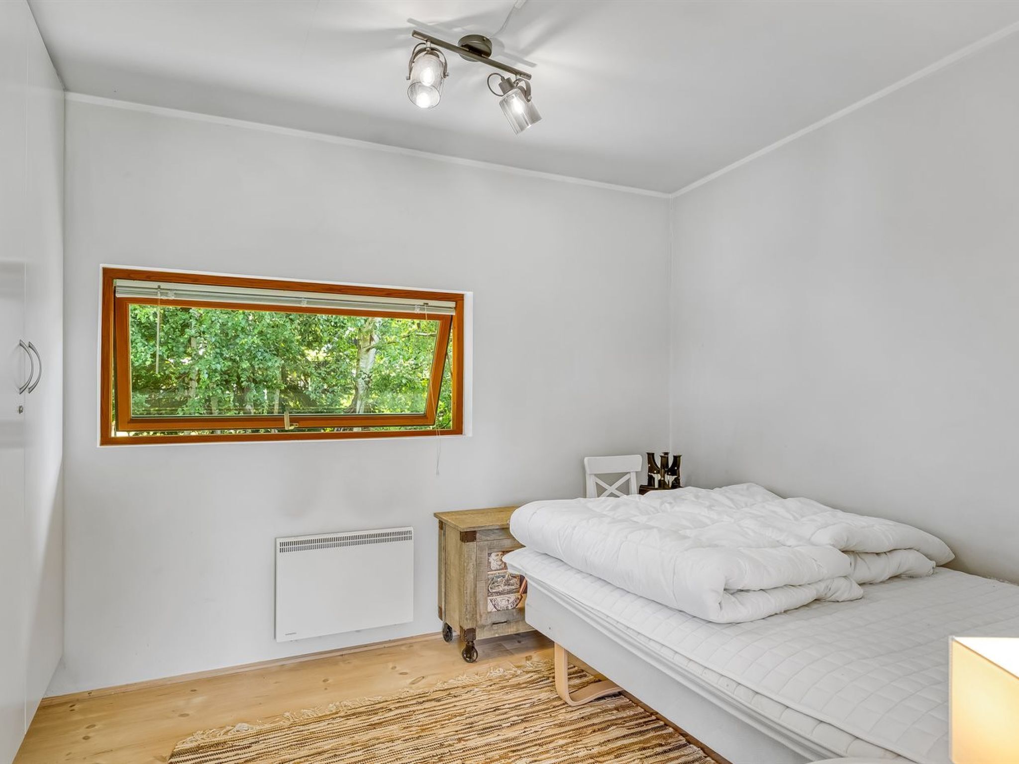A bedroom with a bed, a window with a view of trees, a radiator, and a ceiling light fixture.