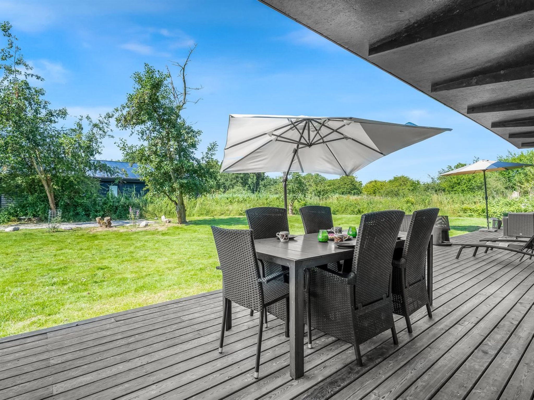 An outdoor patio with a table and chairs, a large umbrella, and a view of a grassy yard with trees.