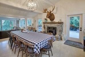 A dining area with a large checkered table, wooden chairs, a stone fireplace, and a mounted deer head on the wall. - Bayfront PNW Lodge: Dock, Pickleball & Guest House (Bainbridge Island)