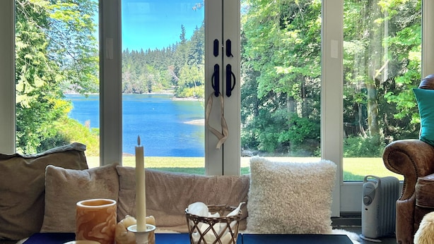 Habitación con un gran ventanal que ofrece vistas a un lago y un bosque. Hay un sofá, una mesa con velas y una silla.