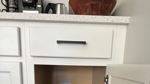 Une cuisine avec des meubles blancs, un plan de travail sur lequel se trouve une machine à café, et une étagère en bois où sont pliés des vêtements et des chaussures.