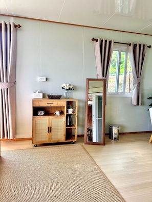 Une pièce avec un meuble en bois, un miroir, une fenêtre avec des rideaux et une petite poubelle.