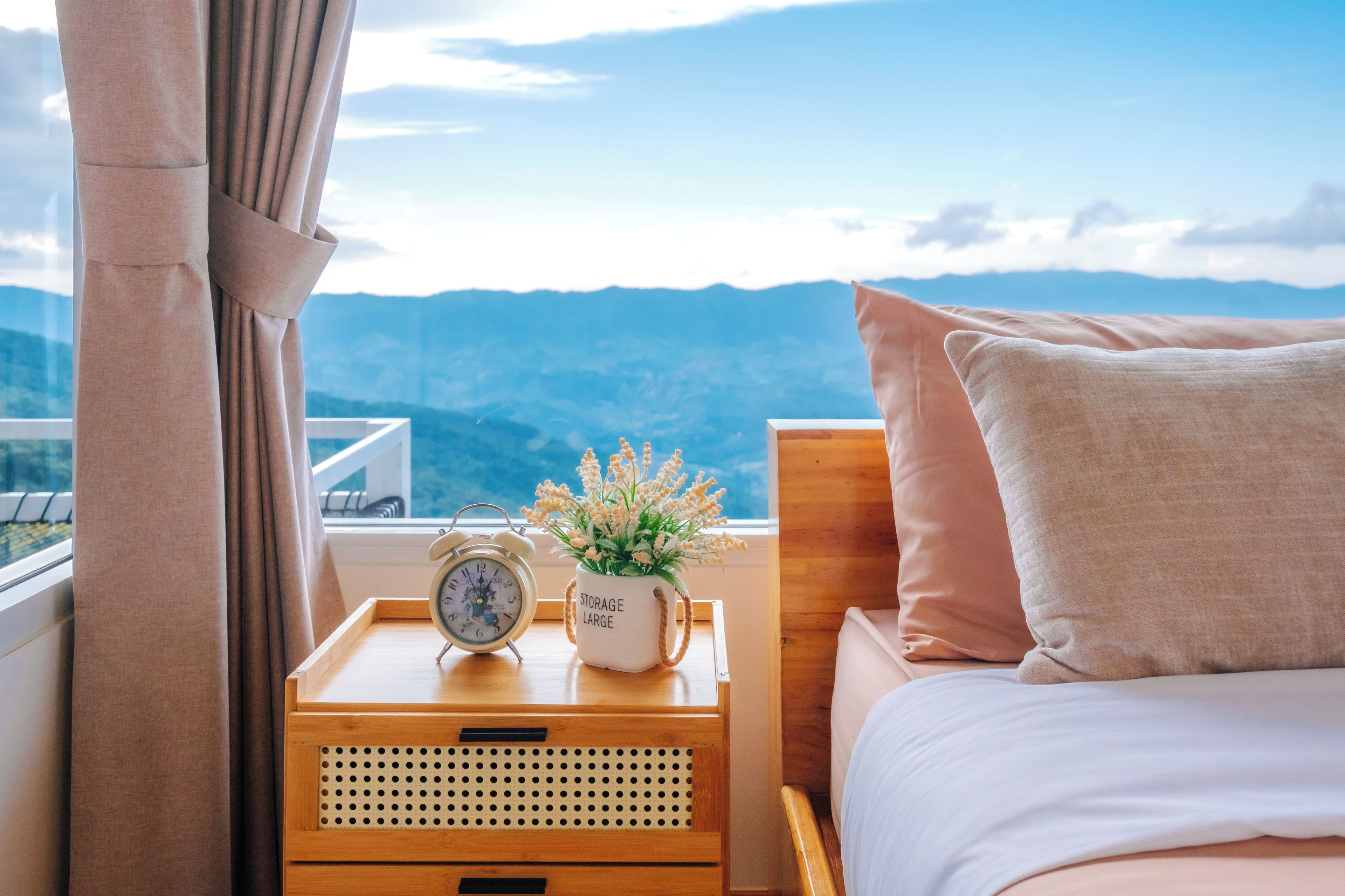 Un dormitorio con una cama, una mesita de noche con un reloj y una planta, y vistas a montañas.