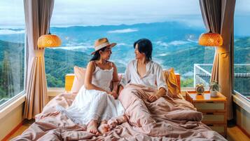 A bedroom with a large bed, two bedside lamps, a nightstand with a clock, and a view of a mountainous landscape.