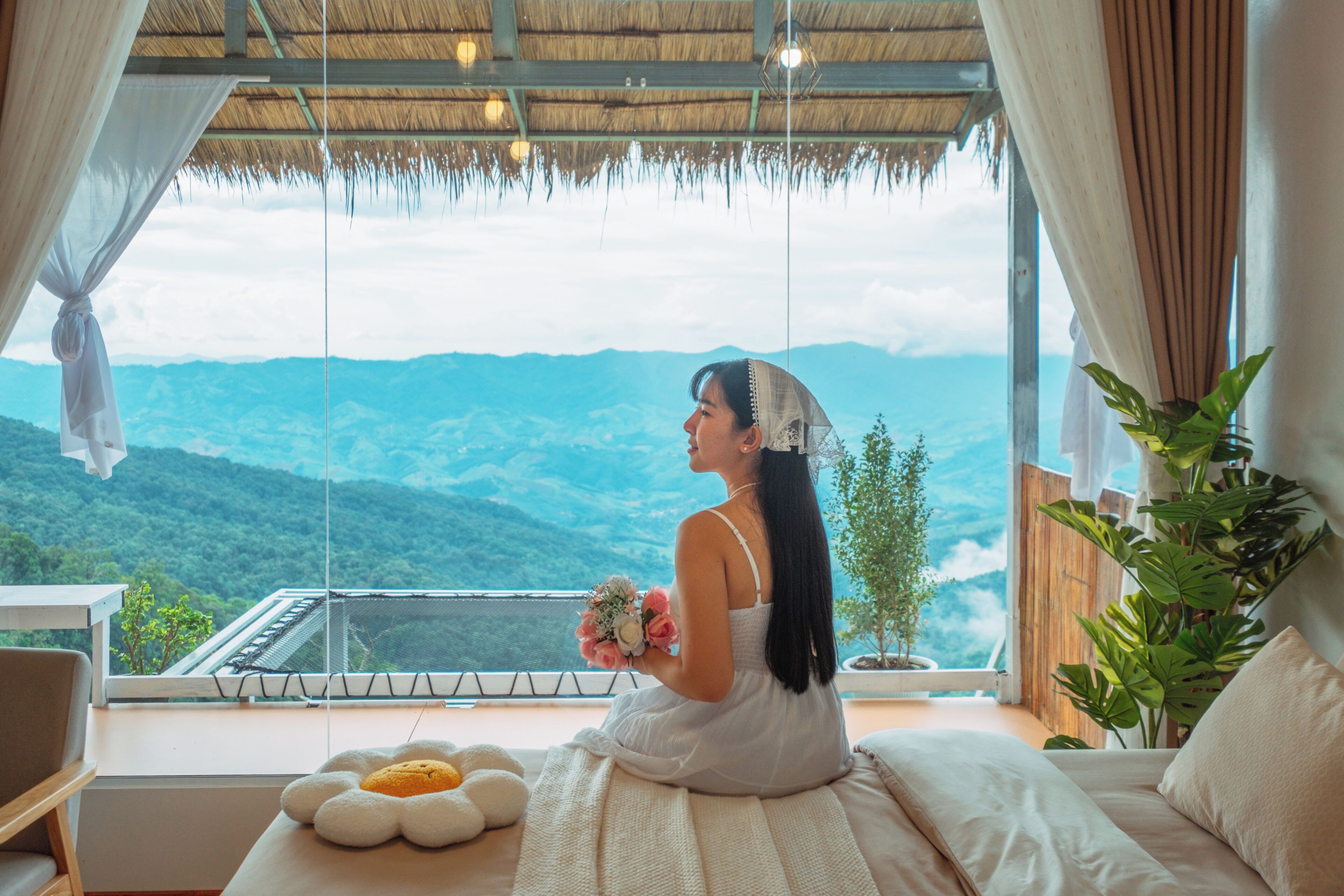 Quarto com uma grande janela com vista para uma paisagem montanhosa, uma cama com almofada florida branca e um vaso com planta.