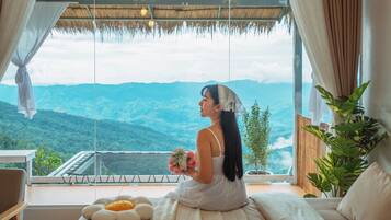 Quarto com uma grande janela com vista para uma paisagem montanhosa, uma cama com almofada florida branca e um vaso com planta.