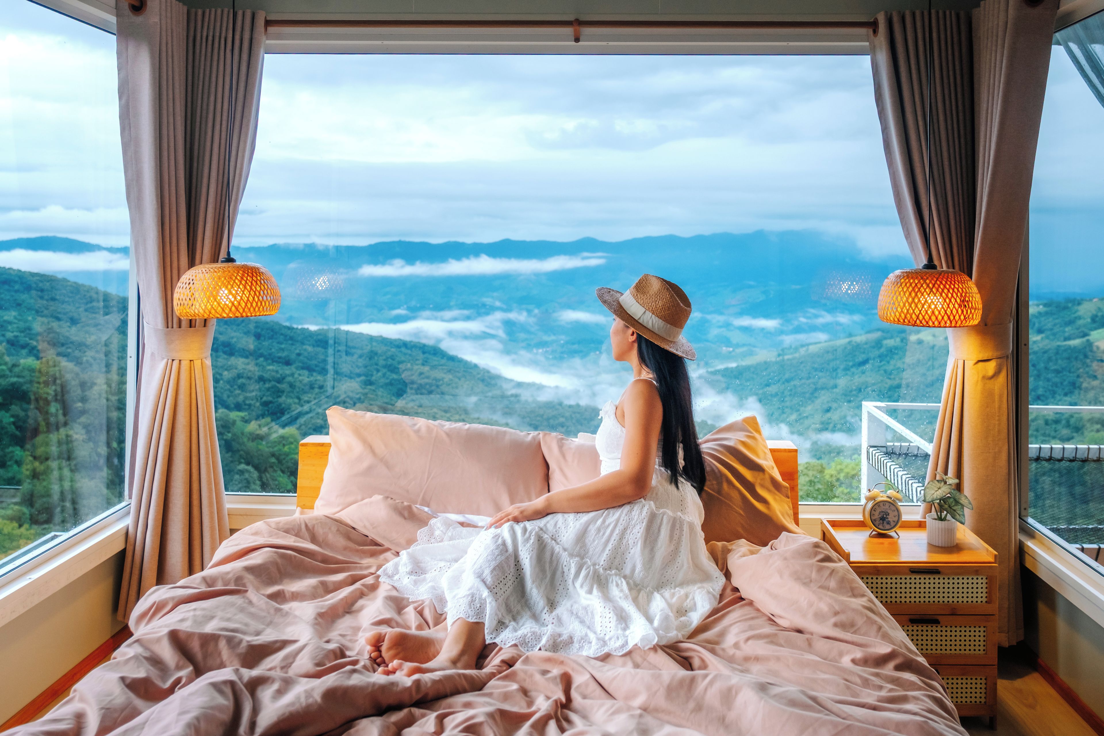 A room with a bed facing a scenic mountain view, a bedside table with a lamp, and a window with curtains.