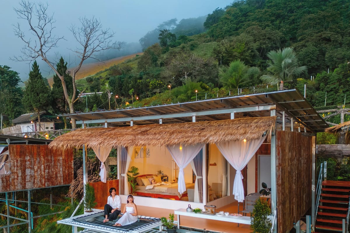 Un hébergement aménagé comme une cabane dans les arbres, avec un toit de chaume, un balcon doté d’une rambarde en métal et une vue sur la forêt environnante.