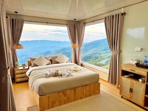 Una camera da letto con un letto grande, un comodino in legno, un cesto di vimini e vista sulle montagne.