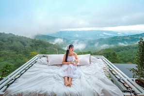 Un letto con biancheria bianca, una persona seduta sopra e vista su un paesaggio montuoso.
