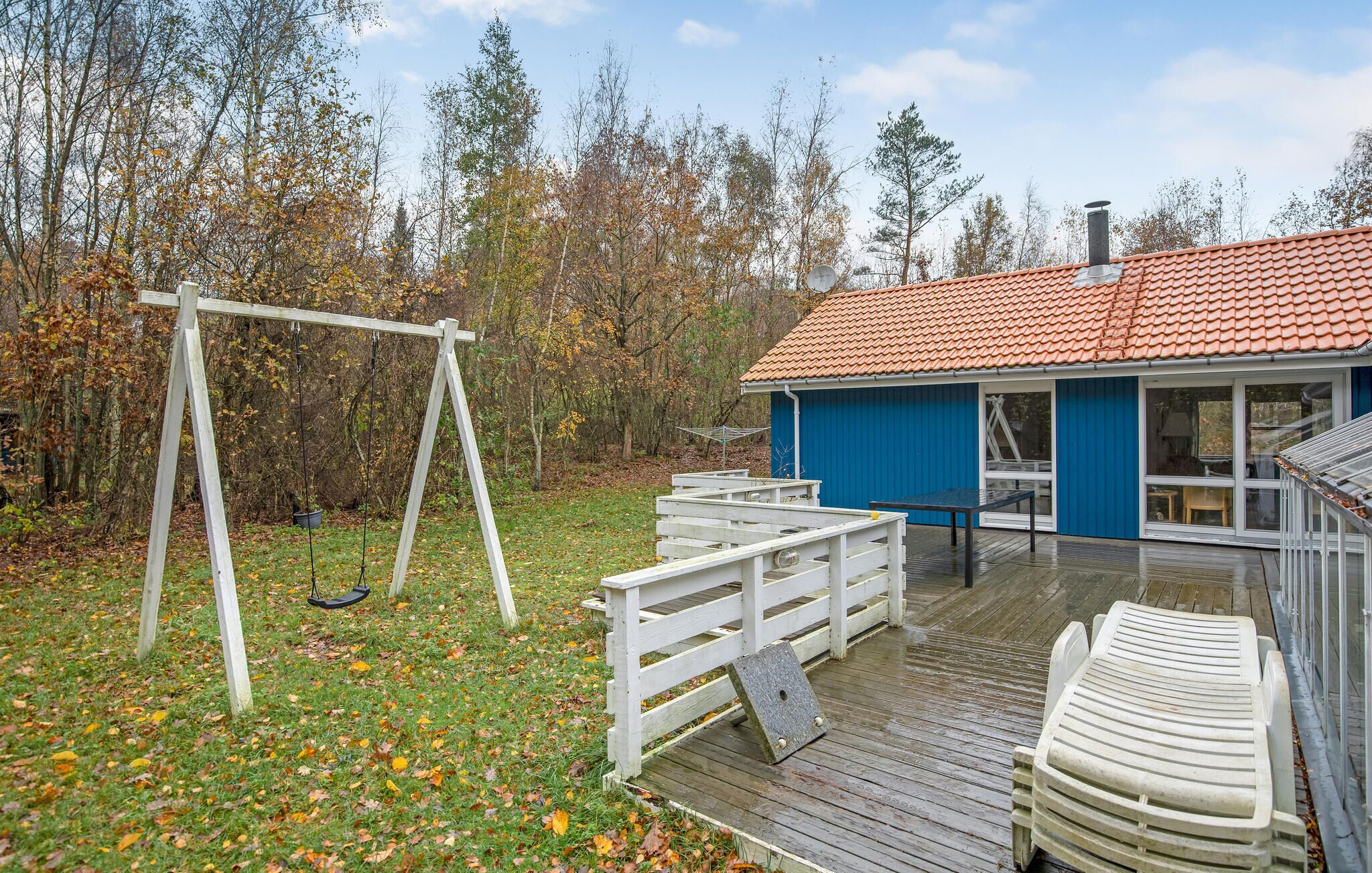 Een houten terras met een schommel, een overdekt buitenverblijf met een tafel en stoelen, en een gebouw met een rode pannendak.