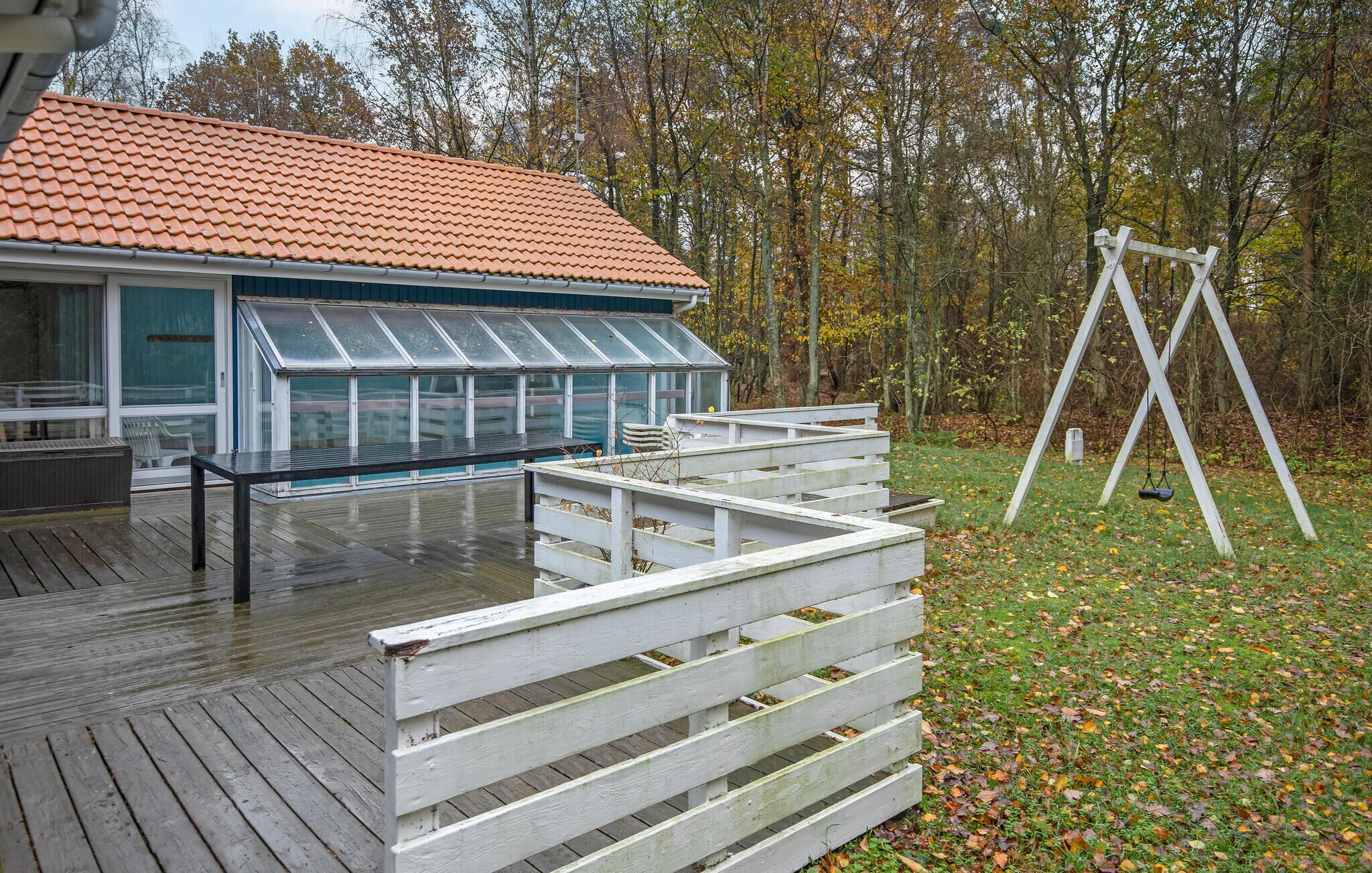Une terrasse en bois avec une pièce fermée par des baies vitrées, une balançoire blanche et un toit recouvert de tuiles rouges.