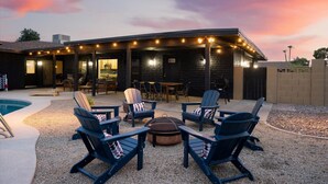 A covered outdoor seating area with Adirondack chairs and a fire pit, surrounded by a pool and a building with a brick exterior. - Sport Court | Private Heated Pool | Mini Golf & More (Glendale)