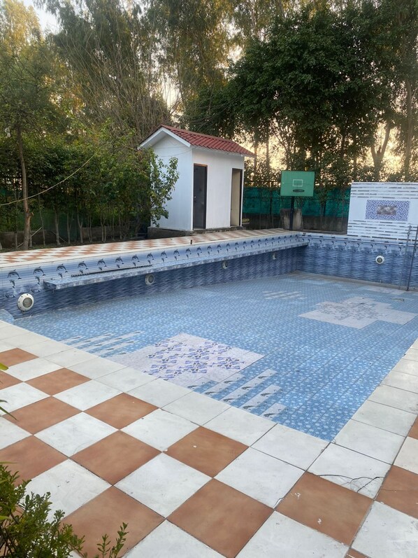 En swimmingpool med fliseterrasse, en lille hvid bygning med rødt tag og et grønt hegn i baggrunden.