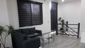 Een moderne woonkamer met een zwarte bank, twee potplanten, een glazen salontafel en een trap met een metalen leuning.