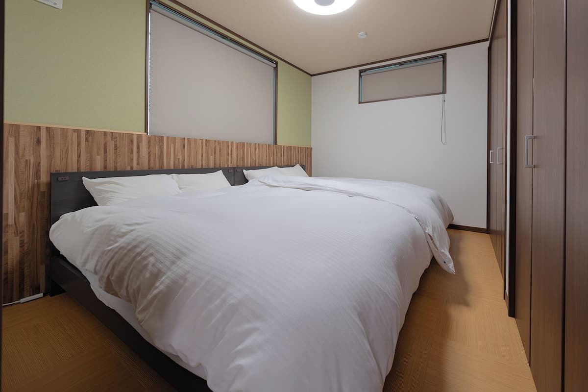 A neatly made bed in a hotel room with a wooden headboard, a small window, and a wardrobe.