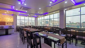 A spacious dining area with multiple tables and chairs, large windows offering a city view, and modern ceiling lighting. - Treebo Empire Red Carpet (Agra)