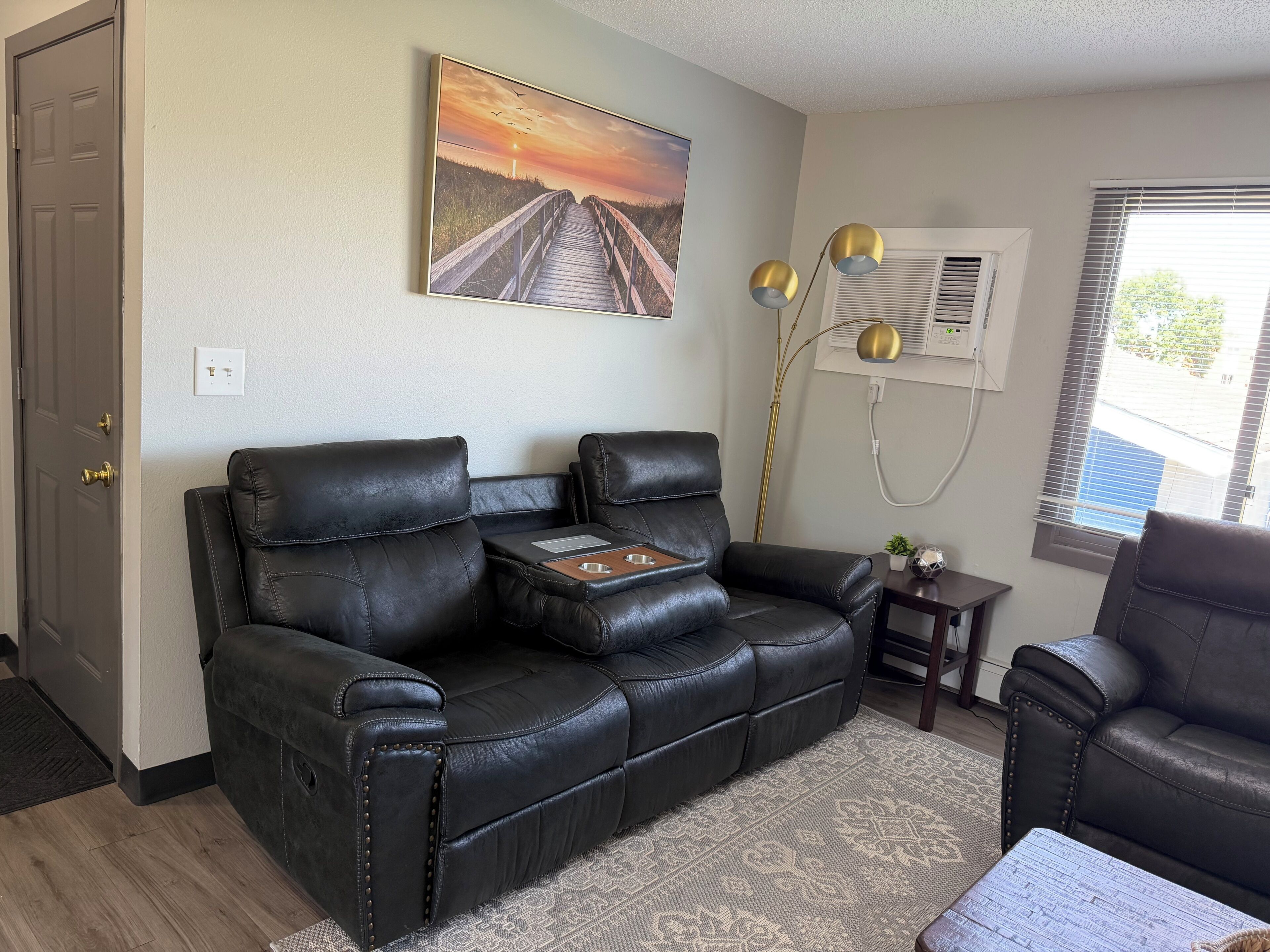 A living room with a black leather sofa, a small side table, a decorative lamp, and a painting of a sunset over a beach.