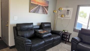 A living room with a black leather sofa, a small side table, a decorative lamp, and a painting of a sunset over a beach.