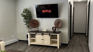 A living room with a flat-screen TV mounted on the wall, a wooden console table with decorative items, and a patterned rug on the floor.