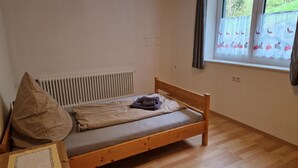 Un llit individual amb estructura de fusta, un radiador, una finestra amb cortines i una presa elèctrica.