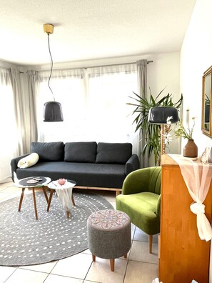 A living room with a dark sofa, a green armchair, a round ottoman, a small table, and a lamp.