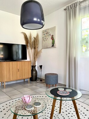 A modern living room with a flat-screen TV, a wooden cabinet, a round glass coffee table with a pink flower centerpiece, and a painting of butterflies on the wall.