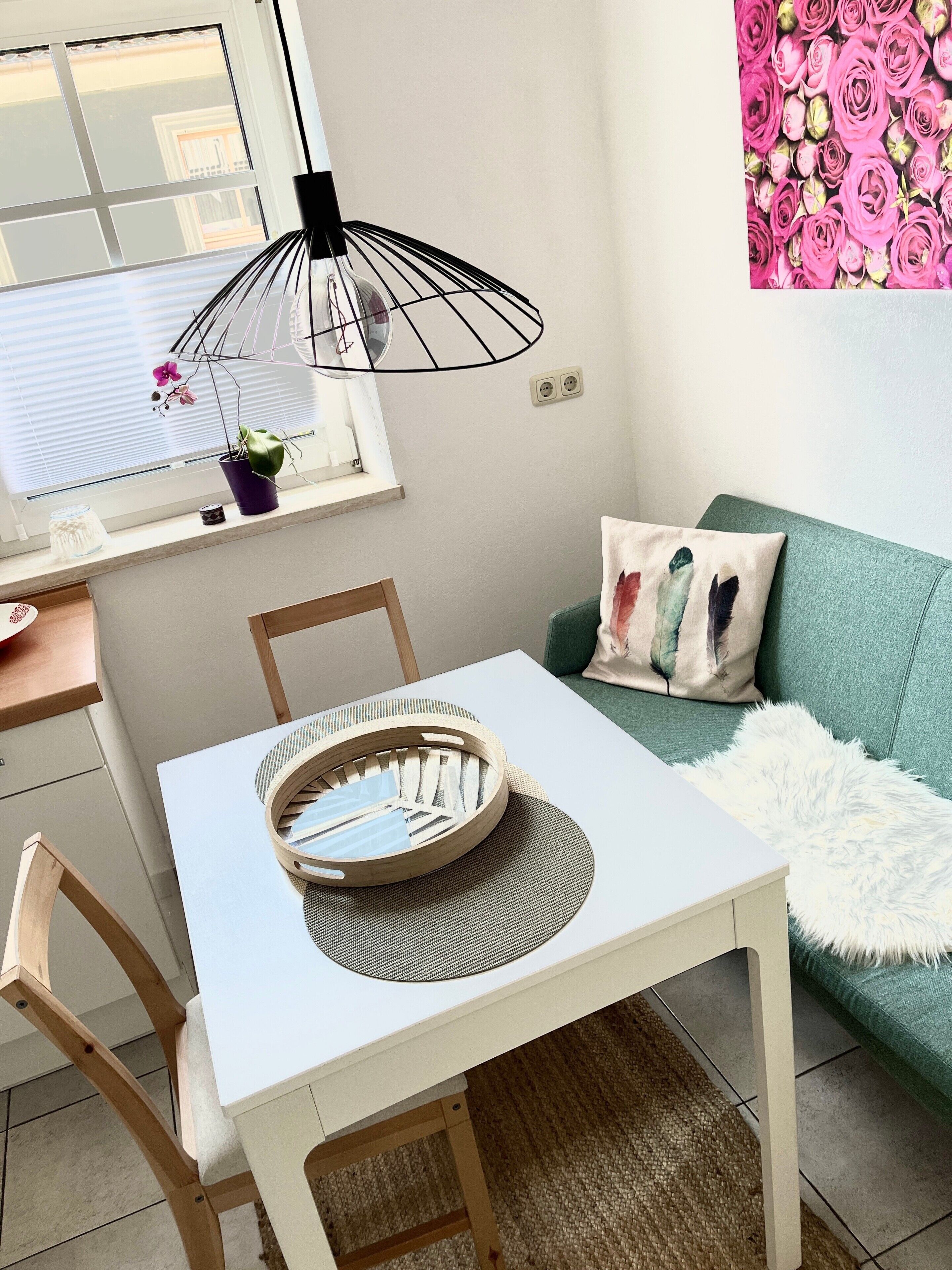 Een eettafel met stoelen, een hanglamp, een raam met jaloezieën, een groene bank met een zachte witte deken en een muur met een bloemenprint.