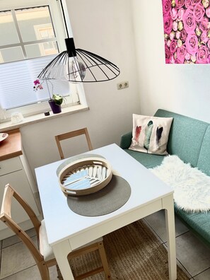 Une table à manger avec des chaises, un luminaire suspendu, une fenêtre avec des stores, un canapé vert agrémenté d’un plaid blanc moelleux et un mur orné d’un motif floral.