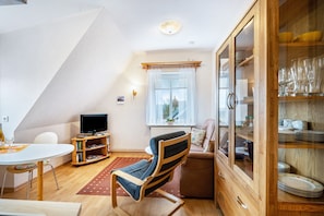 Une pièce comprenant une table à manger, un téléviseur, un fauteuil rembourré, un meuble avec des portes en verre et une fenêtre avec des stores.