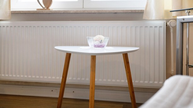 Ein kleiner runder Tisch mit einer klaren Glasbowl, die ein weißes Objekt enthält, sowie Holzbeine und ein weißer Heizkörper im Hintergrund.