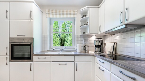 Cocina moderna con armarios blancos, horno empotrado, ventana con vistas a árboles y una campana extractora de acero inoxidable.