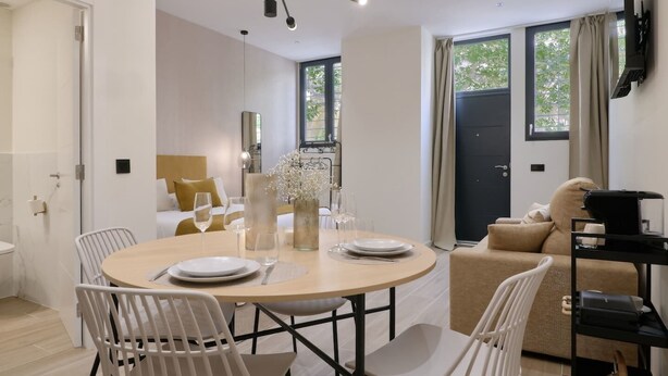 A modern hotel room with a round dining table, white chairs, a bed with a gold headboard, a large window with curtains, and a mounted TV. - Romantic Apartment N5 Oceanografico Virkin Suites (València)