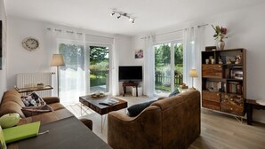 Ein Wohnzimmer mit Sofa, Couchtisch, Fernseher, Bücherregal und Blick durch große Fenster.