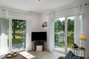 En stue med fjernsyn, lampe og udsigt til en have gennem store vinduer.