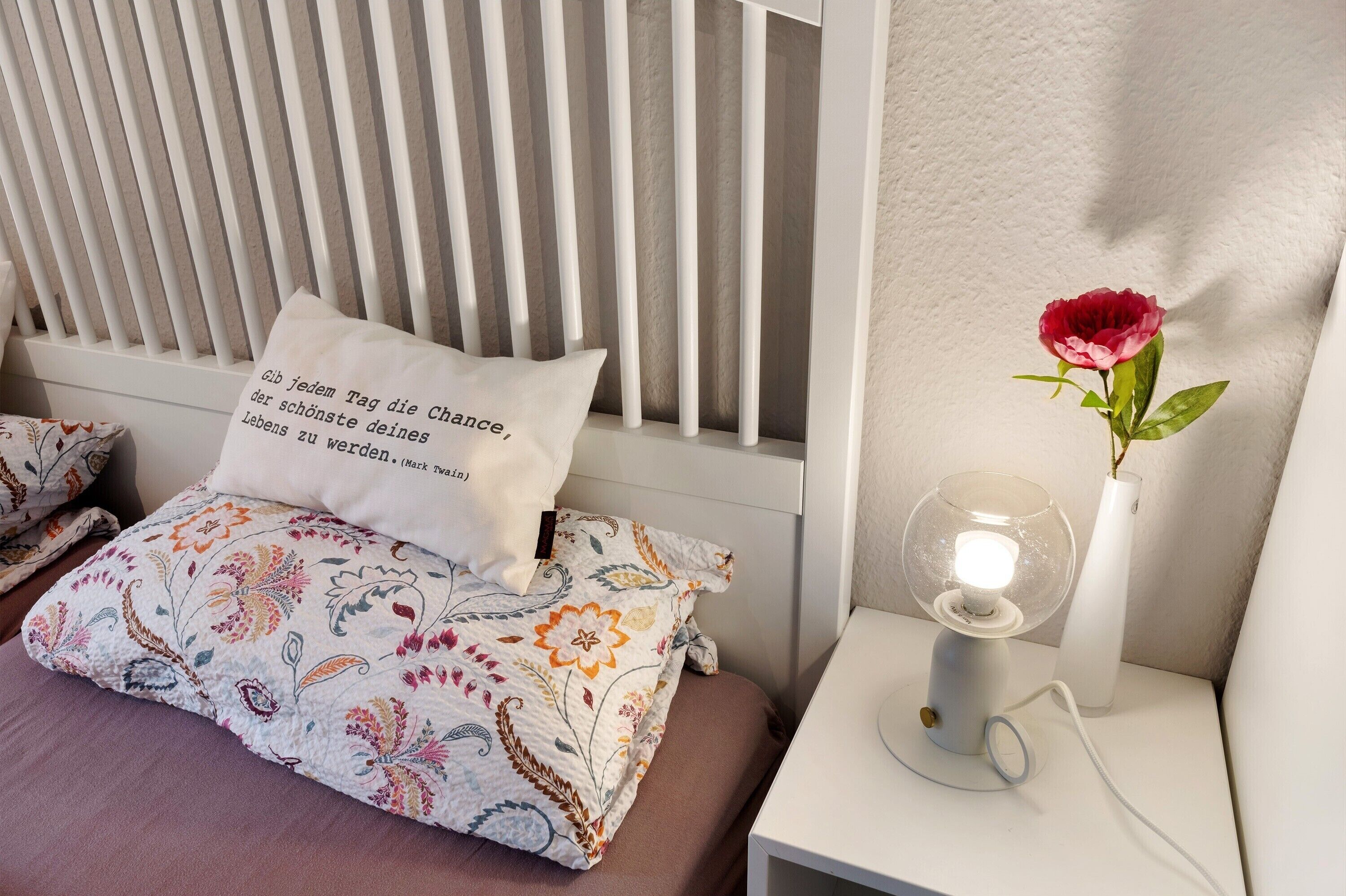 Un dormitorio con una cama, almohadas, una mesita de noche con lámpara y un jarrón con una flor.