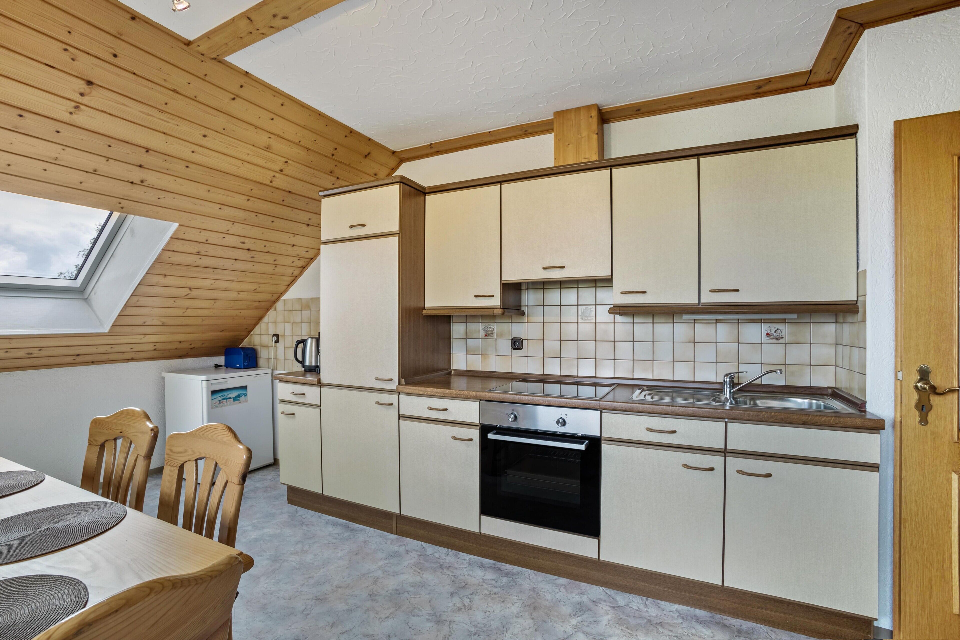 Une cuisine avec des poutres apparentes en bois au plafond, des meubles blancs, un four encastré et une fenêtre.