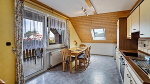 A dining area with a table and chairs, a window with curtains, a radiator, and a kitchen with cabinets and a sink. - Apartment 'Ferienwohnung Bächer' with Shared Garden, Balcony and Wi-Fi (Wiesau)