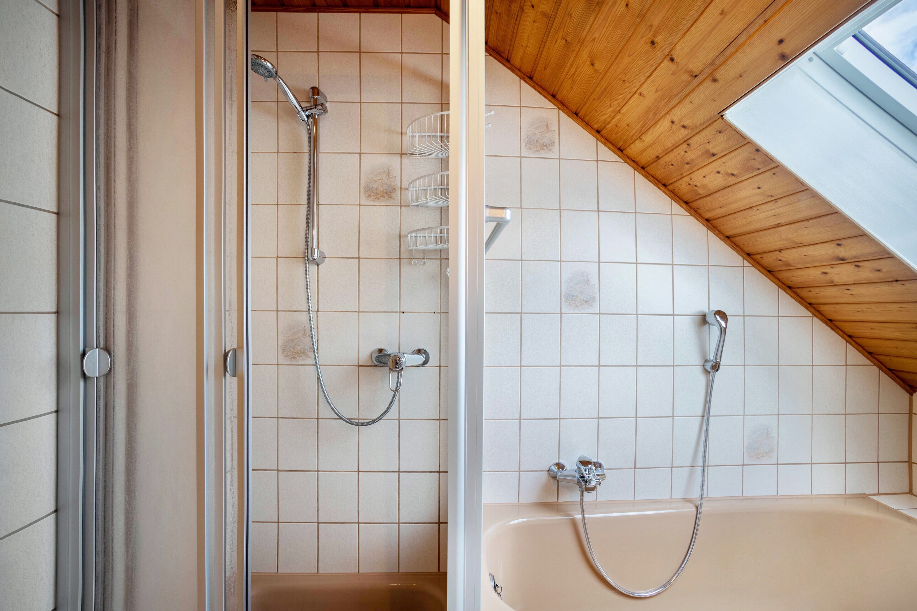 Een badkamer met een douche, een badkuip, betegelde wanden en een schuin houten plafond.