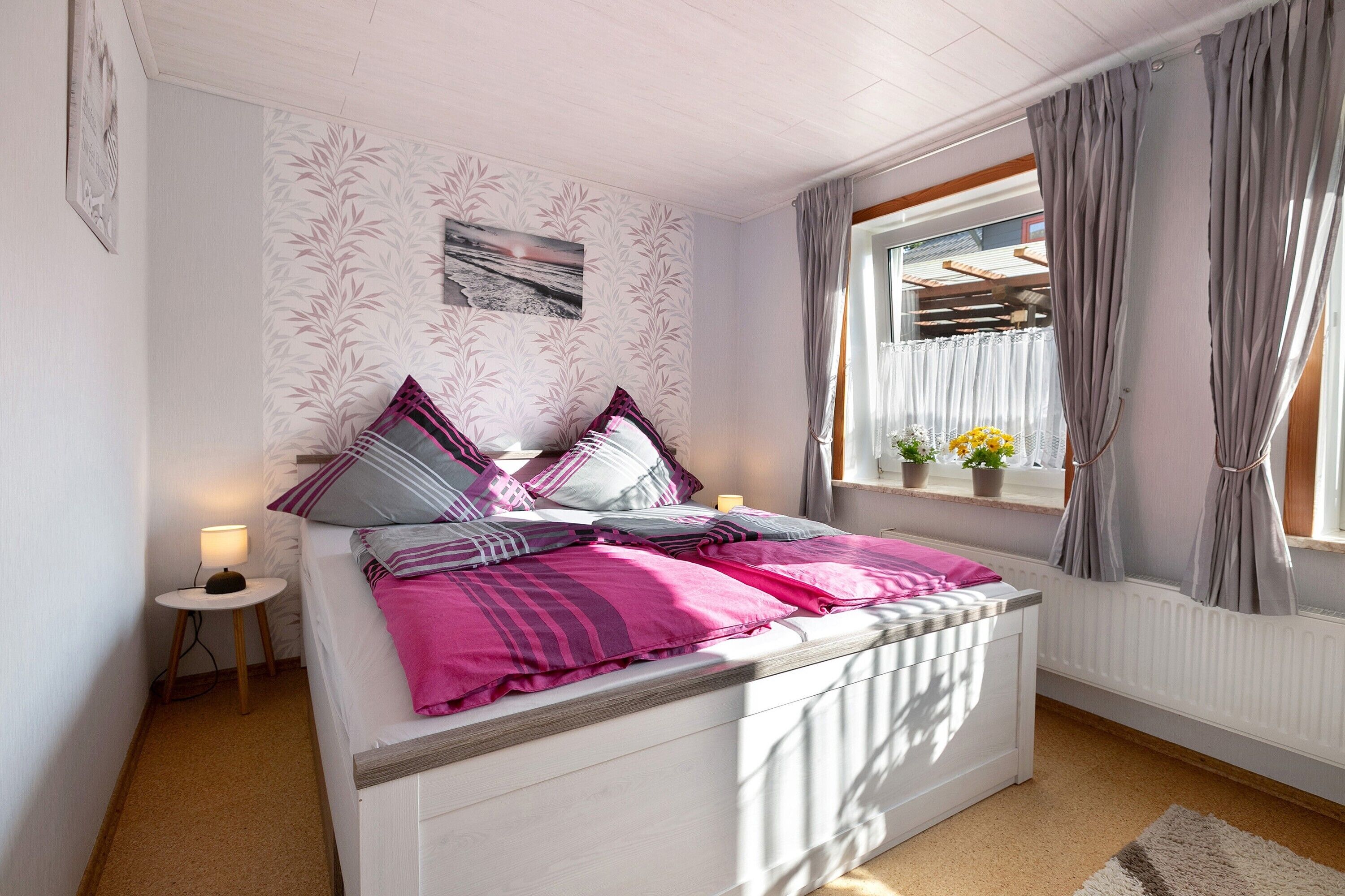 Une chambre à coucher comprenant un lit, une table de chevet, une fenêtre avec des rideaux et un mur orné d’un motif de feuilles.
