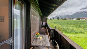 A wooden deck with a table and chairs, a window with curtains, and a view of a green field and mountains. - Apartment 'Ferienwohnung 3' with Mountain View, Shared Garden and Wi-Fi (Grassau)