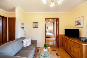 Una sala de estar con un sofá, una mesa de centro de vidrio con flores, un televisor sobre un mueble de madera y un pasillo que conduce a un dormitorio.